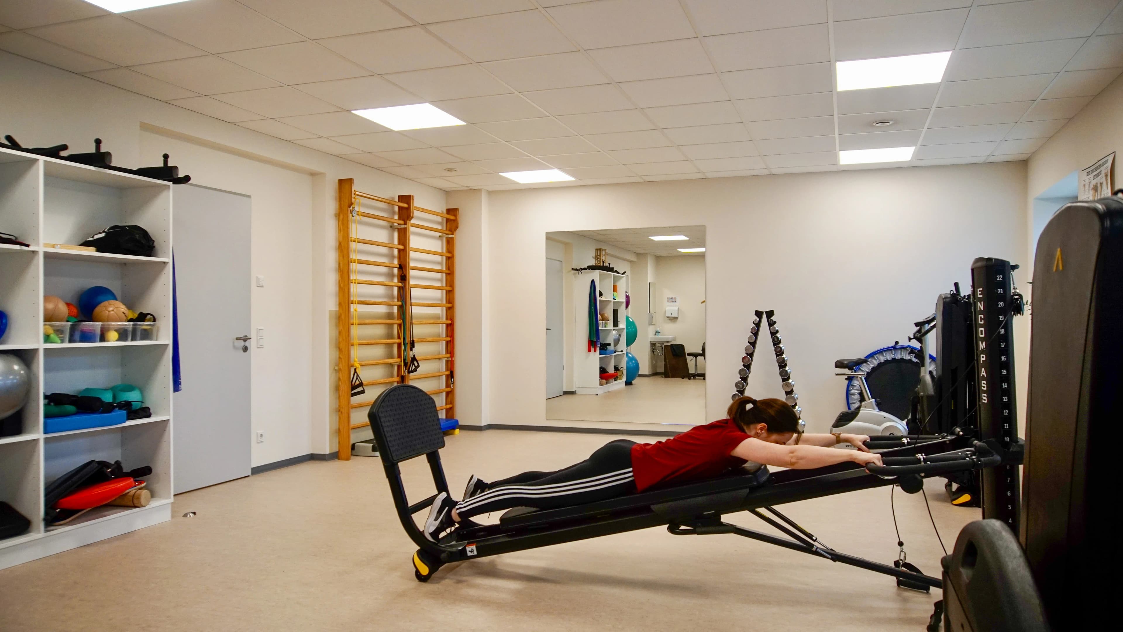 Patientin trainiert an einem Seilzuggerät im Geräteraum mit Sprossenwand, Spiegel und Gymnastikbällen