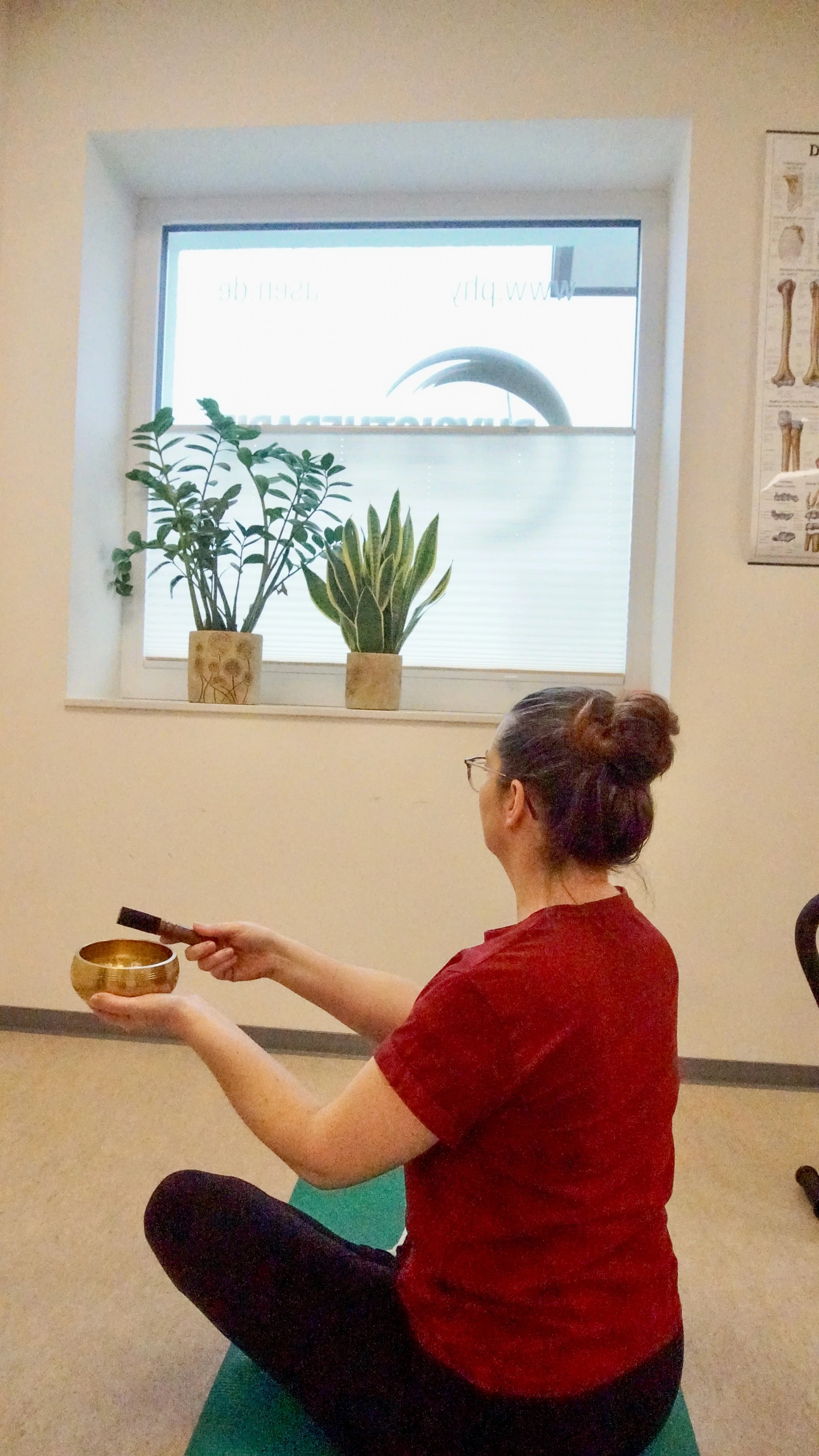 Therapeutin sitzt im Schneidersitz auf einer Matte und spielt eine Klangschale, umgeben von Grünpflanzen